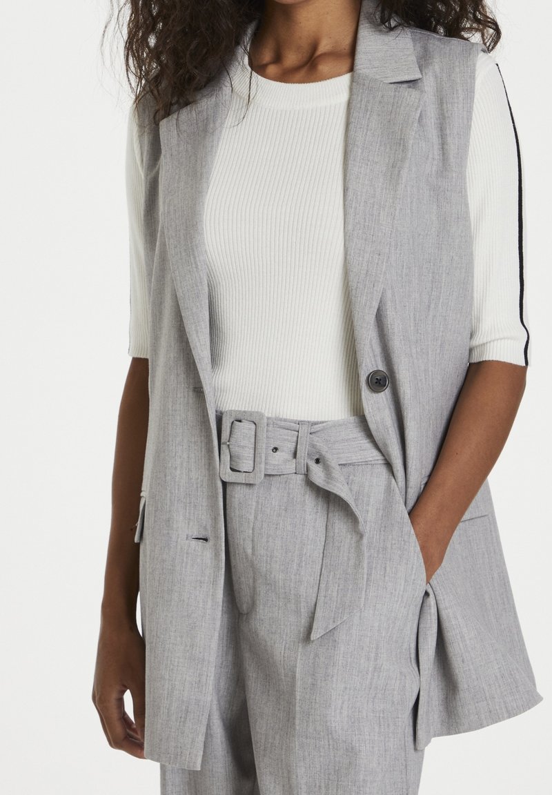 Femme portant un blazer sans manches gris clair et un pantalon ceinturé par-dessus un haut blanc côtelé à manches trois-quarts avec des rayures noires.