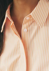 Chemise rayée orange clair avec un col classique, des boutons blancs et une texture de tissu lisse.