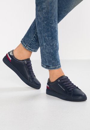 Person wearing dark blue sneakers with red, white, and blue stripes and dark blue laces, paired with blue denim jeans.