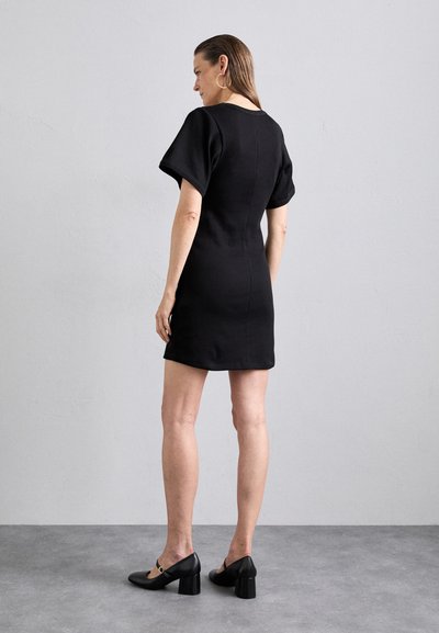 Schwarzes, tailliertes Kleid mit kurzen, weiten Ärmeln. Glattes Material, bis zur Mitte des Oberschenkels und mit einem runden Ausschnitt. Getragen mit schwarzen Absatzschuhen.
