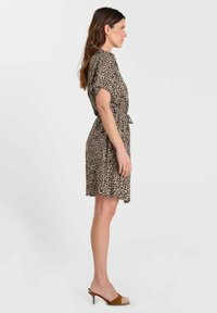 Femme debout de profil portant une robe mi-longue à imprimé léopard avec manches courtes et des mules marron à talons sur un fond uni.