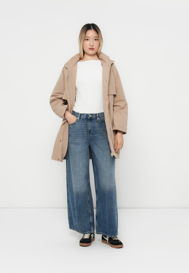 Beżowy, lekki płaszcz z kapturem, zestawiony z szerokimi, ciemnoniebieskimi dżinsami. Pod spodem biała koszula, a całość uzupełniają czarno-beżowe sneakersy.