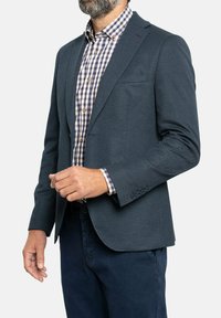 Blazer texturizado azul marino con solapas de muesca, bolsillo en el pecho, y puños de cuatro botones, combinado con una camisa de botones de cuadros debajo.