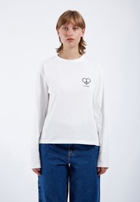 White long-sleeve t-shirt made of cotton, featuring a small black heart logo on the left chest and a relaxed fit paired with blue denim jeans.