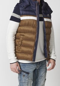 Padded vest with hood, featuring a brown body, navy blue shoulders, and gray striped accents. Zippered front and side pockets.