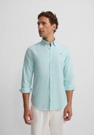 Hombre con camisa de botones de rayas azules claras con las mangas arremangadas y pantalones blancos, mirando hacia su izquierda sobre un fondo liso.