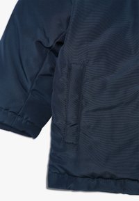 Doudoune bleu marine au tissu lisse, avec une poche latérale et des poignets élastiques à l'ourlet. Détails de couture visibles.