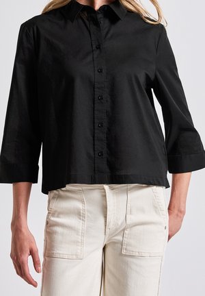 Kvinde iført en sort skjorte med knapper og trekvartærmer og højtaljede, off-white bukser med store forlommer.