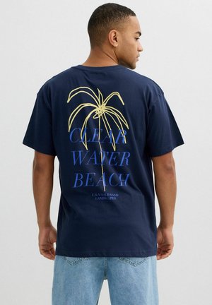 Hombre con camiseta azul marino con un gráfico amarillo de palmera y texto azul "CLEAR WATER BEACH" en la parte trasera, combinado con jeans azul claro.