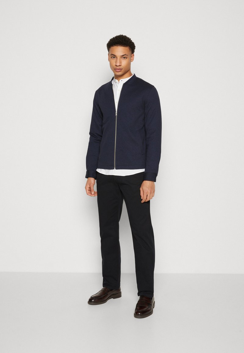 Veste zippée marine en tissu tricoté avec un col de type bomber, portée sur une chemise blanche, associée à un pantalon noir et des chaussures marron.