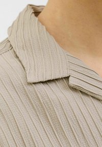 Beige overhemd met kraag, voorzien van een verticale geribbelde structuur en een subtiele glans. De kraag heeft een ontspannen vorm, wat het ontwerp als geheel versterkt.