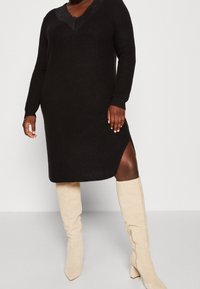 Žena v černých pletených midi šatech s krajkovým výstřihem a bočním rozparkem, ve spojení s béžovými semišovými vysokými kozačkami, na jednobarevném pozadí.