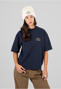 Camiseta de algodón azul marino de manga corta, con un logo bordado en dorado. Combinada con pantalones color caqui y un gorro crema de canalé.