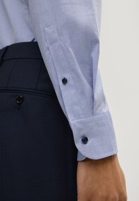 Maniche di camicia a righe blu chiaro con polsino abbottonato, abbinate a pantaloni blu scuro. Presenta bottoni a contrasto e una texture liscia.
