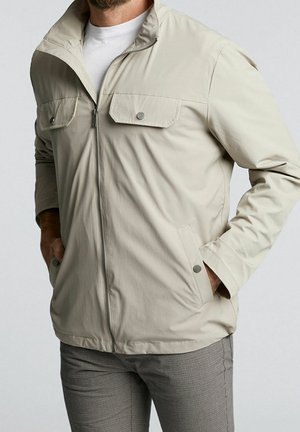 Mann trägt eine hellbeige Reißverschlussjacke mit Brustklappen und die Hände in den Seitentaschen, kombiniert mit grau karierten Hosen.