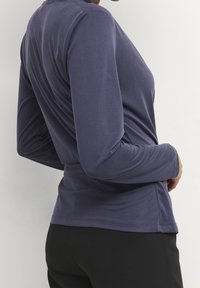 Person i en åtsittande, lång ärmad mörkblå topp och svarta byxor, vänd sidan mot en enkel ljus bakgrund.