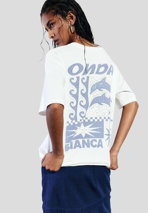 White t-shirt with blue graphic design on back featuring dolphins and patterns. Model wears a dark skirt, with wet hair and neutral background.