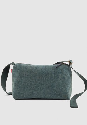 Majhen pravokotni denim crossbody nahrbtnik z nastavljivim temno zelenim trakom in zadrgo na enobarvni podlagi.