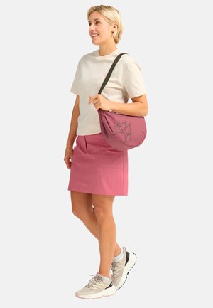 Frau steht, trägt ein beiges T-Shirt, einen rosa Rock, beige Turnschuhe und trägt eine rosa Umhängetasche mit schwarzem Pfotenabdruck-Design.
