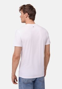 Weißes Baumwoll-T-Shirt mit kurzen Ärmeln und rundem Ausschnitt. Verfügt über ein kleines Logo-Detail am oberen Rücken. Kombiniert mit blauen Jeans.