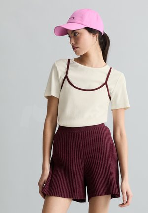 Jonge vrouw met een roze pet, een crèmekleurige top met korte mouwen en bordeauxrode biezen, en bordeauxrode geribbelde gebreide shorts, die opzij kijkt.