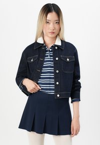 Denimjakke i mørkeblå med sølvknapper og lommer med klaff, kombinert med en poloskjorte i marineblå og hvite striper og en plisséskjørt i marineblå.