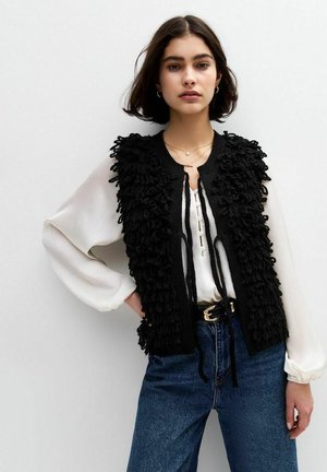 Svart stickad väst med texturerad, fluffig detalj, parat med en vit blus med knappkrage och elastiska ärmar, bärs tillsammans med denimjeans.
