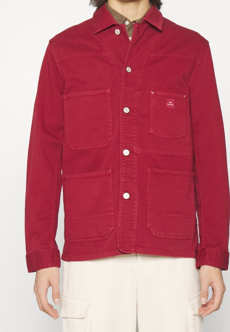 Veste en coton rouge avec un devant à boutons, un col pointu et quatre poches avant. Les détails remarquables incluent des boutons et des coutures contrastants.