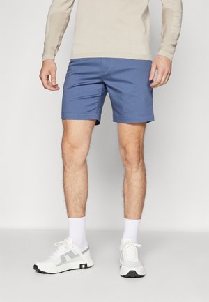 Shorts albastre dintr-un material ușor, cu un design simplu, având o partea din față și un tiv netede, asortați cu adidași și șosete albe.