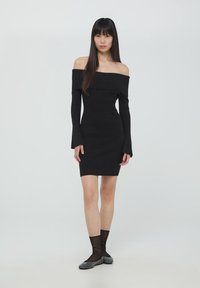 Abito nero a costine con scollo a spalle scoperte, maniche lunghe, design aderente e orlo che termina sopra il ginocchio. Abbinato a calze nere trasparenti e ballerine con motivo.