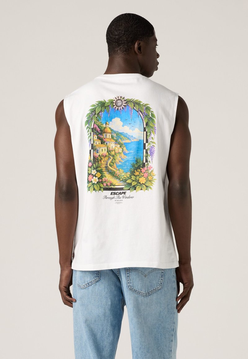 Man draagt een wit mouwloos shirt met een kleurrijke kustdorp- en oceaandruk, omlijst door bloemen en groen op de achterkant.
