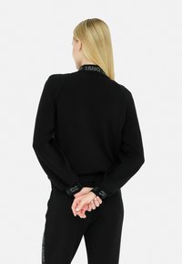 Sweatshirt noir avec un col côtelé, des manches longues et des accents de logo sur les poignets. Le tissu semble lisse et légèrement texturé.
