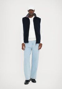 Veste en polaire marine avec deux poches avant, superposée à un sweat-shirt blanc. Un pantalon large bleu clair et des chaussures noires complètent la tenue.