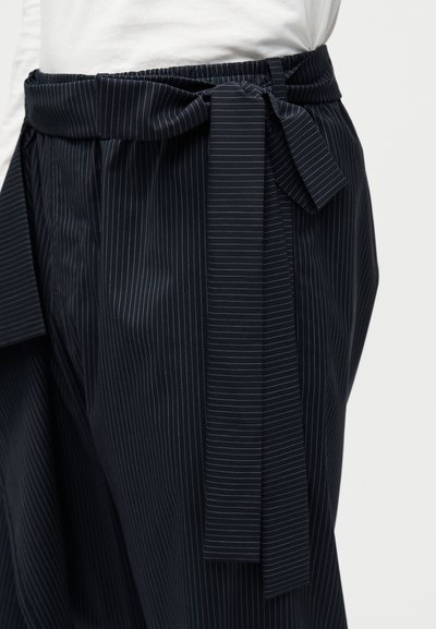 Κοντινό πλάνο σε σκούρα φούστα με λεπτές ρίγες και δέσιμο από ύφασμα στη μέση, πάνω από λευκό τοπ, αναδεικνύοντας τη μέση και τη λεπτομέρεια της ζώνης.