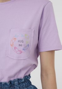 T-shirt corto viola con maniche corte, caratterizzato da una tasca con mani multicolori disposte in un design circolare e il testo "ABBRACCIAMI" al centro.