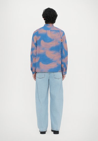 Long-sleeve shirt featuring a blue and peach cloud pattern, semi-sheer fabric, spread collar, and button cuffs, paired with light blue jeans.