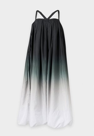 Frau steht, trägt ein lockeres, ärmelloses Ombre-Maxikleid mit dunklem Oberteil, das zum hellen Saum übergeht, und schwarze Sandalen vor einem schlichten Hintergrund.