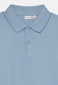 Jasnoniebieska koszulka polo z prążkowanym kołnierzykiem, zapinana na dwa guziki, o gładkiej teksturze materiału. Metka wskazuje rozmiar 9-10 lat, marki o nazwie "name it".