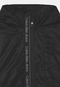 Zwarte hoodie-jack met een gladde textuur, ritsluiting en contrasterend logo aan de voorkant, gepresenteerd in een minimalistisch ontwerp.