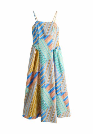 Kobieta stoi na drewnianym tarasie przy basenie, ubrana w bezrękawnikową, wielokolorową, pasiastą, patchworkową sukienkę maxi z cienkimi ramiączkami i sandałami.