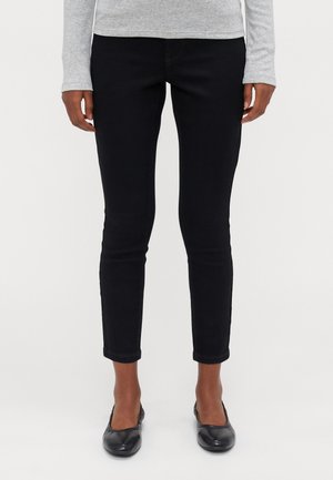 Persoon draagt strakke zwarte enkellange broek, grijze lange mouwen top en zwarte platte schoenen tegen een effen witte achtergrond.