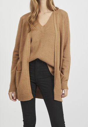 Femme portant un pull col V beige et un cardigan ouvert assorti avec des poches, associé à un pantalon noir taille haute sur un fond uni.