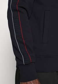 Sudadera navy con tejido texturizado, presenta franjas rojas y blancas a lo largo de la manga y un bolsillo lateral. Tejido suave, puños ajustados.
