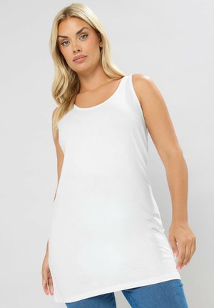 Donna bionda con tunica bianca senza maniche e jeans blu, in piedi davanti a uno sfondo grigio uniforme.