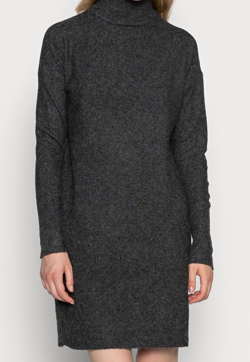 Femme portant une robe en maille à col roulé, manches longues, de couleur gris foncé, debout devant un fond clair uni.