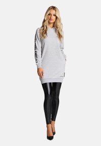 Graues Kapuzensweatkleid mit langen Ärmeln, versehen mit schwarzem Text am Arm, kombiniert mit glänzenden schwarzen Leggings und hohen Absätzen.