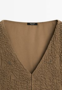 Braunes, strukturiertes Kleid mit V-Ausschnitt, das über geraffte Stoffdetails und kleine dekorative Verzierungen in der Nähe der Schulter verfügt.