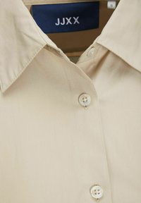 Camicia beige con colletto, bottoni bianchi e etichetta del marchio "JJXX" con la taglia S all'interno del colletto.