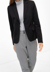 Femme portant un blazer noir sur un col roulé gris clair et un pantalon boutonné à motif pied-de-poule, debout devant un fond blanc.