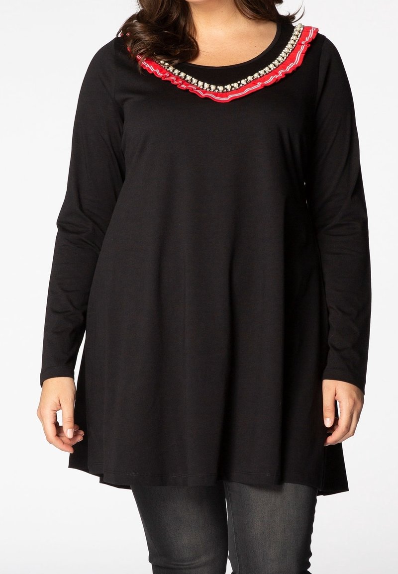 Femme portant une tunique noire à manches longues avec des volants rouges et des détails en perles autour du cou, associée à un jean gris foncé.
